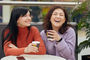 Picture of beautiful emotional young ladies sitting at table in cafe, drinking coffee, holding paper cups, laughing sincerely, having funny talk, wearing casual clothes. Women and friendship concept. clipart