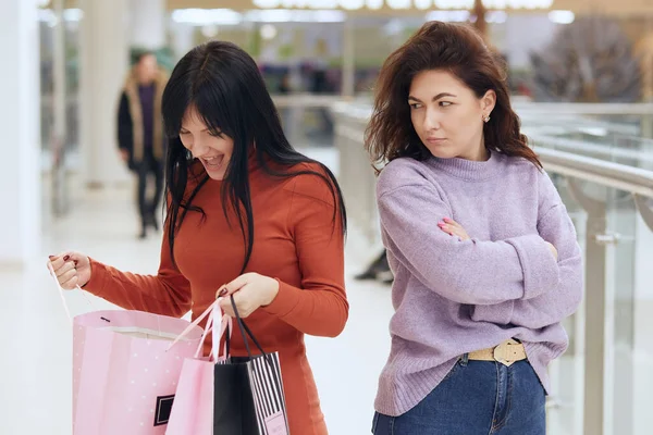 Horizontal shot of two beautiful charming young women standing in shopping centre. Brunette wearing orange dress, holding packages with purchases, looking inside. Her friend is offended at female.