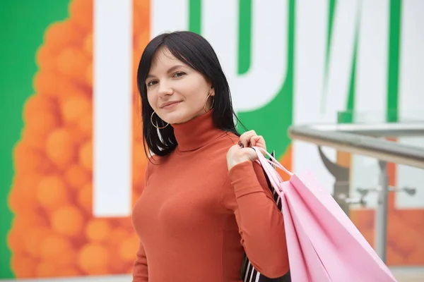 Horizontal shot of woman holding shopping bags and smiling at directly at camera in shopping mall, dark haired adorable female wearing orange casual sweater, doing shopping and has positive emotions.