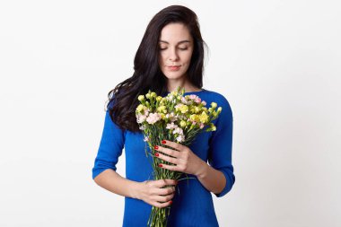 Koyu renk saçlı genç bir kadının portresi. Beyaz arka planda izole edilmiş çiçek demetleri tutuyor ve kokluyor. Esmer kadın, mavi elbiseli, gözleri kapalı poz veriyor. Halk konsepti,