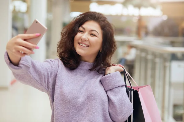 Indoor shotof beautiful girl with shopping bags and taking selfie with their cell phone, looking smiling at device\'s screen, wearing purple stylish sweater, walks in shopping mall. Lifestyle concept.