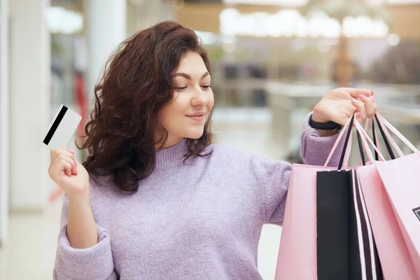 一方で買い物袋と別のクレジットカードで幸せなブルネットの女性の水平ショットは 彼女の新しい服を見て ショッピングモールにポーズをとって 喜びを表現し 幸せそうに見えます ライフスタイルコンセプト — ストック写真