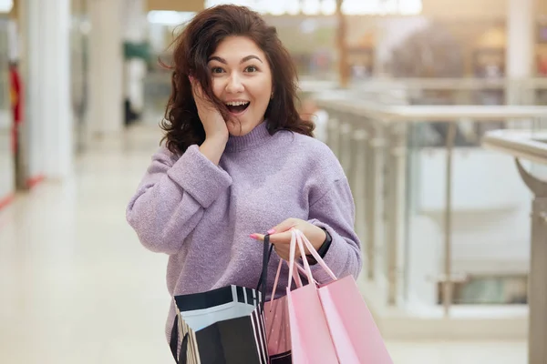 ショッピングバッグを持つ幸せなショッピング女性の水平方向のショットは 驚きの表情を持つカメラを見て ショックを受け 彼女の頬に手のひらを維持し スタイリッシュな服を着ている ショッピングのコンセプト — ストック写真