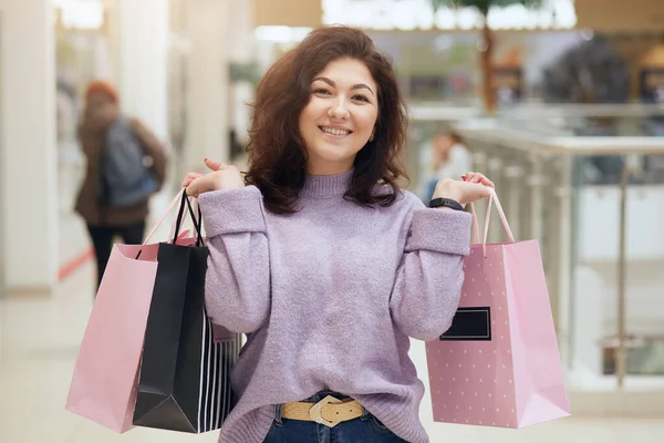 ショッピングモール 愛らしいブルネットのドレスライラックセーターを歩いている間 カメラを見て笑顔で買い物袋を持っている美しい女の子の肖像画を閉じ 幸福を表現しています ショッピングのコンセプト — ストック写真