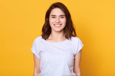 Sarı arka planda yalnız poz veren beyaz bir kadın, kameraya doğrudan mutlu bakan büyüleyici gülüşlü bir kadın, siyah saçlı sevimli bir kız, güzel haberler veren, mutluluğunu ifade ediyor. İnsanların duyguları