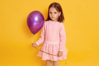 Kapalı alanda, pembe balon tutan, pembe elbiseli, sarı arka planda tek başına duran, kameraya bakan güzel, güzel, güzel, sarışın küçük bir kızın resmi. Çocukluk kavramı.