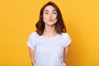 Sarı arka planda izole edilmiş beyaz tişört giyen güzel, koyu renk saçlı bir kadının portresi. Bir kadın kameraya bakıyor, havada öpüşüyor..