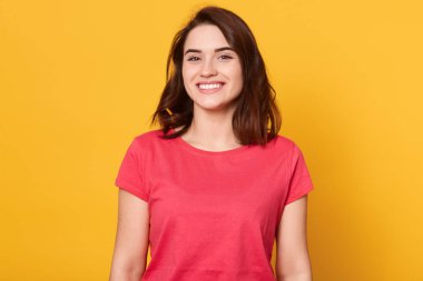 Siyah saçlı çekici bir kadının görüntüsü çok mutlu, geniş bir gülümseme ile mükemmel dişlerini gösteriyor, kırmızı günlük tişört giyen heyecanlı bir kadın, sarı arka planda izole edilmiş poz veriyor..