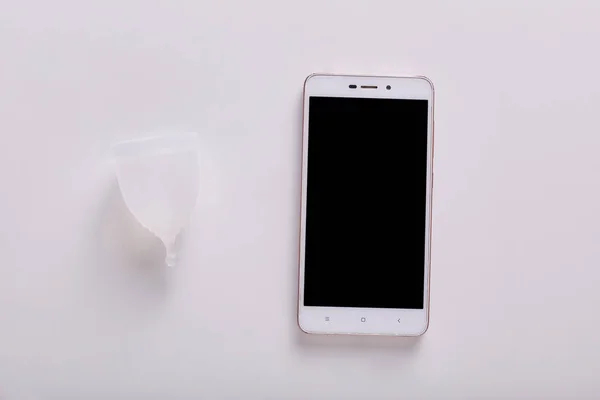 Innenaufnahme von sauberer, weißer Menstruationstasse aus Gummi, die auf weißer Oberfläche in der Nähe von Smartphone mit leerem Bildschirm liegt, installiertem Zeitrahmen, alternativer umweltfreundlicher Typ. Frauen und Menstruationskonzept. — Stockbild Innenaufnahme Von Sauberer Weißer Menstruationstasse Aus Gummi Die Auf Weißer — Stockfoto