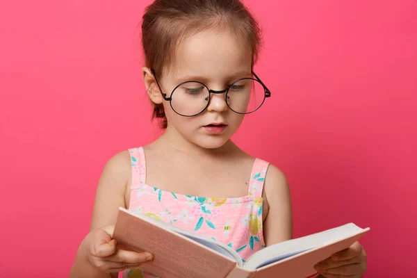 穿着夏装和眼镜的可爱小女孩 手里拿着书 神魂颠倒 读着有趣的童话 诗歌或故事 靠着粉红的画室墙壁站着 儿童教育概念 — 图库照片
