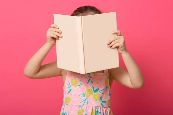 在玫瑰色的工作室背景下 女孩躲在书本后面的横向照片 穿着裙子的女孩喜欢阅读有趣的童话或故事 在学校有任务 — 图库照片