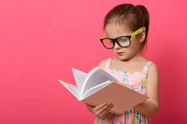 照片上 可爱的学龄前女孩穿着太阳衣和眼罩 女孩独立站在玫瑰工作室的背景上阅读课本 孩子们喜欢读有趣的童话 — 图库照片