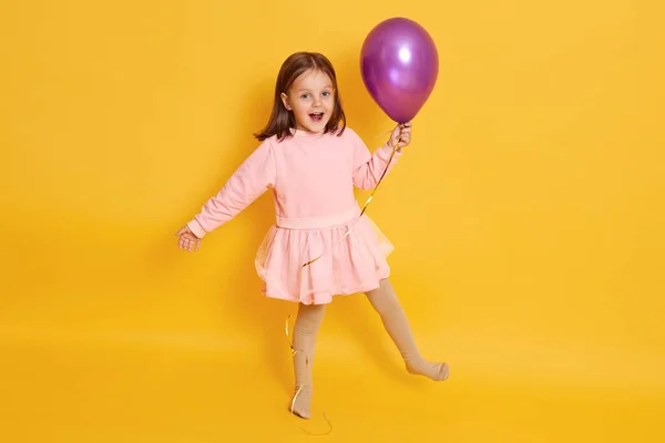 Studio shot of joyful adorable cute little child wearing light pink dress, holding balloon, opening mouth and eyes widely, standing on one leg, playing game, spending time alone, having fun.
