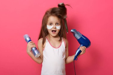 Pembe stüdyo arka planında yalnız poz veren sevimli bayan çocuk portresi, küçük bir kız saçını fırçayla tarıyor, sorunlar yaşıyor, saç modelini yaparken birbirine dolanmış, saç kurutma makinesi kullanıyor..