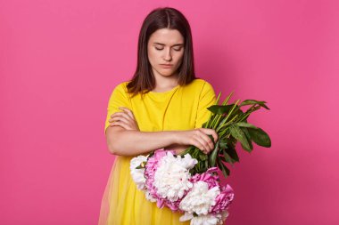 Pembe stüdyo arka planında izole edilmiş çiçekleri tutan güzel esmer bir kadın, sarı elbiseli bir kadın, katlanmış elleri olan koyu saçlı bir kadın, gücenmiş görünüyor, erkek arkadaşıyla tartışıyor..