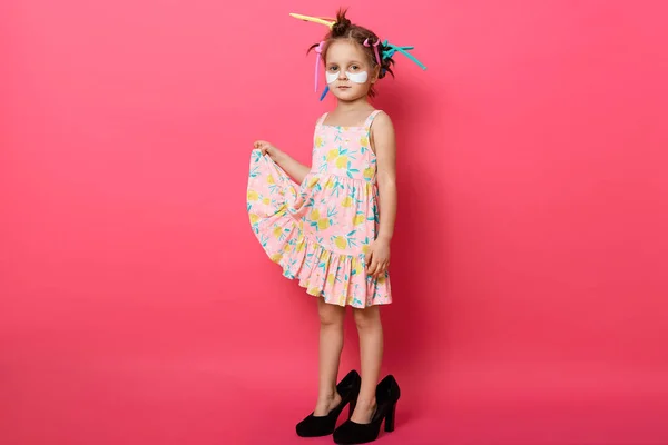Horizontal shot of little girl wearing big mothers shoes and sundress, posing with hair curlers, cute kid standing with patches under eyes isolated over rose background. Child fashion concept.