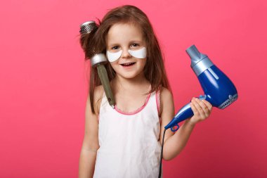 Gül stüdyosunun arka planında izole edilmiş, elinde mavi saç kurutma makinesiyle poz veren sevimli bir kız çocuğu, saçlarını taramış, kafasında saç fırçası olan küçük bir kız. Çocuk konsepti.