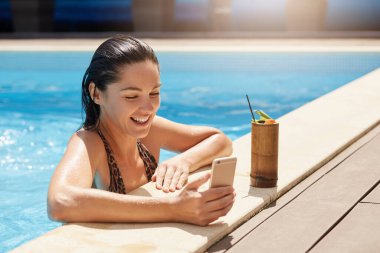Sevimli, neşeli, tatlı bir kadının yatay görüntüsü samimi bir şekilde gülümsüyor, selfie çekiyor, sosyal ağ siteleri için fotoğraf çekiyor, akıllı telefon tutuyor, havuzda yüzüyor, ahşap bardakta kokteyl içiyor..