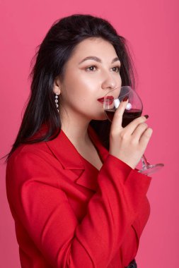 Dikey çekimde, beyaz güzel bir kadının kırmızı şık bir elbise giyip şarap kadehi tutarken, zevkini çıkarırken, bakışlarını kaçırırken, sevimli esmer kadın pembe stüdyo duvarına poz verirken..