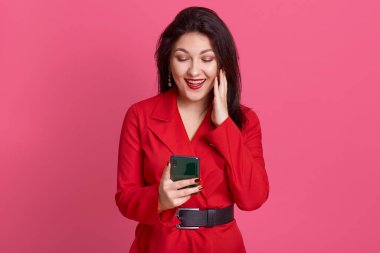 Kırmızı ruj sürmüş kırmızı elbiseli mutlu genç bir kadın cep telefonuna bakıyor ve gülümsüyor. Heyecanlı bir yüz ifadesi var. Pembe arka planda izole edilmiş poz veriyor..