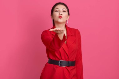 Güzel bir esmerin, hava öpücüğü gönderen, elini kaldıran, jest yapan, kameraya doğrudan bakan şık kemer ve kırmızı ceket giyen güzel bir esmerin kapalı mekândaki portresi. İnsanlar ve duygular.