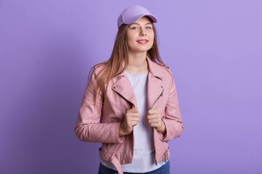 Pembe deri ceket, beyaz gömlek ve kep giyen genç ve modaya uygun modelin stüdyo portresi. Kadınlar moda ve yaşam tarzı konsepti. Leylak stüdyosunun arka planında kendine güvenen kadın pozları