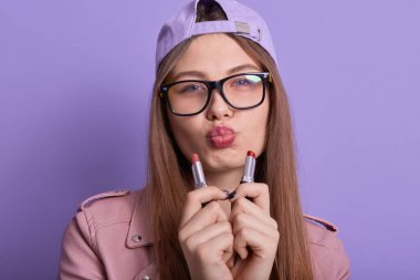 Çok çekici bir kadının, kameraya doğrudan bakan portresini yakın çekimde, iki elinde ruj tutarken, seçim yaparken, güzellik ürünleri yaparken, modaya uygun aksesuarlar giyerken. Güzellik konsepti.