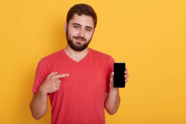 Akıllı telefonu elinde tutan ve işaret parmağı sarı arka planda izole edilmiş siyah ekranı işaret eden bir adamın kapalı çekim görüntüsü. Kırmızı tişört giyen ve telefonunu gösteren sakallı adam. Boşluğu kopyala.