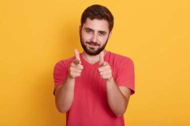 Kırmızı tişört giyen, kendine güvenen, kameraya iki eliyle bakan, sarı arka planda tek başına duran yakışıklı sakallı adamın yakın plan portresi..