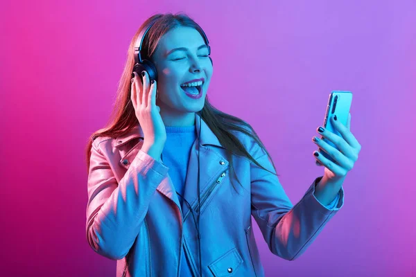 音楽に耳を傾け 片手にスマートフォンを持ち ビデオを録画し 口を広く開き 喜びを持って歌っているヘッドフォンを持つ感情的な美しい精力的な若い女性 テクノロジー コンセプト — ストック写真