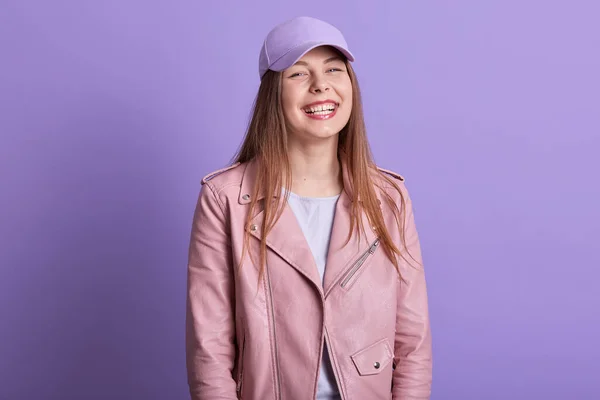 Horizontal shot of fashion woman with long fair hair posing in studio against lilac wall, lady dressedrosy leather jacket and cap, female standing with toothy smile and happy facial expression.