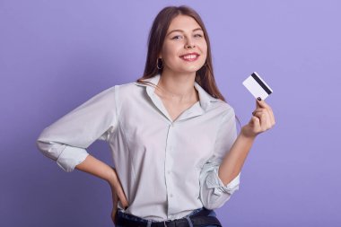 Uzun sarı saçlı, güzel bir kadının portresi, kredi kartı tutuyor, belini tutuyor, doğrudan kameraya bakıyor, günlük kıyafetler giyiyor, güzel bir gülümsemesi var. Para kavramı.