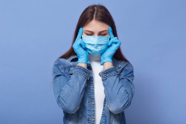 Kafasına dokunan eldiven ve tıbbi maske takan, mavi stüdyo arka planında izole edilmiş, baş ağrısı çeken, şakaklarına dokunan kadın portresi. Corona virüsü, covid 19, sağlık hizmeti konsepti