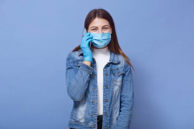 Mavi flüt maskeli, mavi maskeli, beyaz gömlekli ve kot ceketli genç bir kadının koronavirüsten korumaya çalıştığı, arkadaşıyla telefonda konuştuğu yatay bir fotoğraf. Covid 19 konsepti.