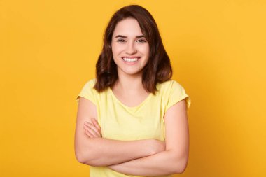 Sarı arka planda izole edilmiş, katlanmış elleri olan güzel gülümseyen bir kadının stüdyo fotoğrafı mutlu görünüyor, neşe veriyor, günlük kıyafetler giyiyor, koyu renk saçlı. İnsan duyguları kavramı.