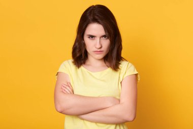 Güzel, koyu renk saçlı, sıradan bir tişört giyen bir kadının portresini yakın çekim. Somurtkan dudaklarıyla kameraya bakıyor, ellerini katlıyor, sarı arka planda tek başına duruyor ve rencide oluyor.