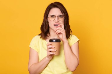 Hoşnutsuz, siyah saçlı, yüzünde hoşnutsuzluk ifadesi olan, elinde kahvesini tutarken parmağını ısıran, üzgün, günlük giysiler giymiş beyaz bir kadın..