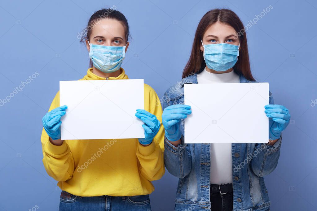 Image of two young women wearing medical disposable masks and medical gloves holding white blank papers in hands, copy space for some inscriptions. Corona virus, covid 19, health care concept.