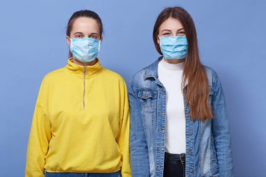 Günlük kıyafet ve sağlık maskesi takan, grip, koronavirüs ya da bulaşıcı hastalıkları önlemeye çalışan mavi arka planda izole edilmiş iki kadının portresi..