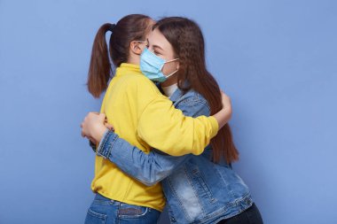 Corona virüsünün yayılmasını önlemek için günlük giysiler ve koruyucu maskeler takan iki genç kadının stüdyo fotoğrafları. Mavi arka planda birbirlerine sarılan kadınlar..