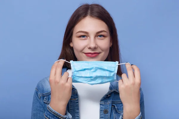 Horizontal shot of beautiful woman puts on medical mask isolated over blue background, girl trying to avoid flu or coronavirus. Female wearing denim jacket with flu mask in hands. Healthy care concept