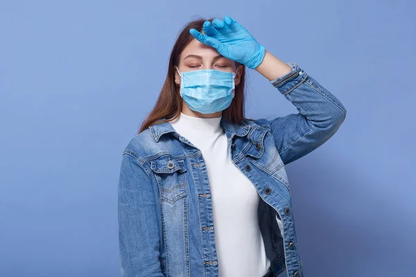 照片中疲惫的年轻女性有发烧 一只手放在额头上 感觉不适 闭上眼睛 头戴胶布 戴着抗菌面具和手套 Coronavirus概念 — 图库照片