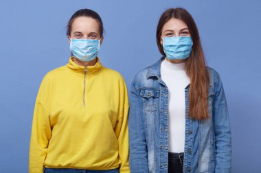 Tıbbi maske takan, kameraya bakan, mavi arka planda izole edilmiş iki genç kadının stüdyo portresi. Grip salgını, toz alerjisi, virüse karşı koruma, koronavirüs konsepti..