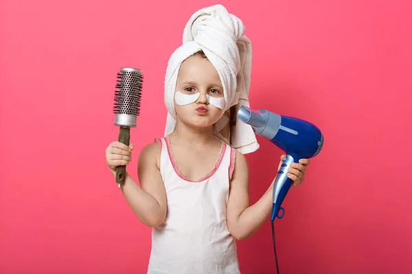 Picture of charming cute female kid isolated over rosy studio studio wall, girl being ready combing her hair with brush, making hairstyle, using hairdryer, adorable little lady keeping lips rounded.