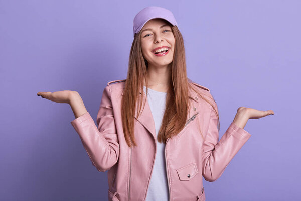 Horizontal shot of smiling teen girl with spreading hands aside, expressing happyness, posing isolated over lilac background, female wearing pink leather jacket, white shirt and cap. People concept.