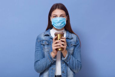 Beyaz ceketli, beyaz gömlekli, mavi koruyucu maskeli, elinde kahve tutan ve kameraya bakan genç beyaz kadın portresi. Coronavirus. covid 19 kavramı.