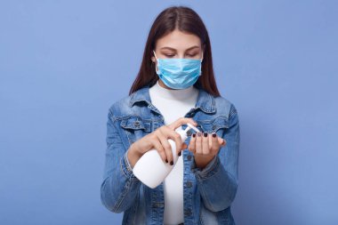 Salgın sırasında sıvı sabun spreyi kullanan bir kadın, beyaz gömlek, kot ceket ve mavi, tek kullanımlık yüz maskesi giyen bir kadın. Coranavirus, covid 19, karantina konsepti.