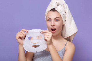 Kafasında beyaz havluyla ve mükemmel cildiyle büyüleyici güzel bir kadının portresi, eflatun stüdyosunun arka planında izole edilmiş kumaş maskesini çıkarıyor. Kozmetik ve cilt bakımı prosedürleri kavramı.
