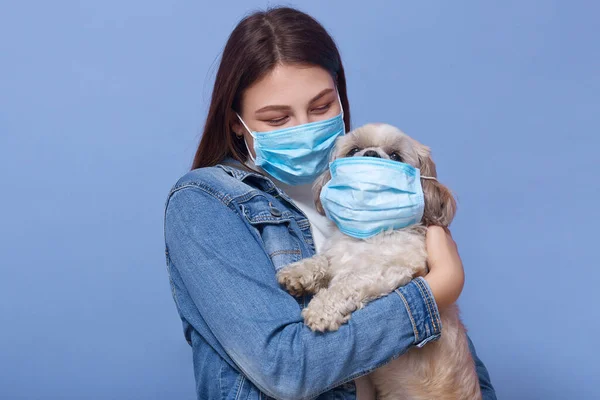 Coronavirus, protection, health, covid 19 and quarantine concept. Young adult caucasian woman and her dog wearing blue madicalprotective face masks, standing isolated over studio background.