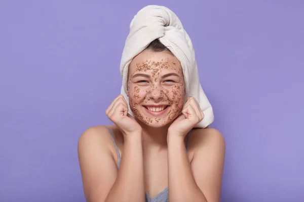 Headshot of smiling happy dark skinned female model has rejuvenation procedure, cleans face with white crystal peeling mask after taking shower, looks younger, isolated over pink background.